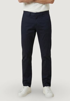 Homme portant un pantalon slim-fit bleu marine foncé, des baskets blanches et un t-shirt blanc, debout bras croisés devant un fond uni.