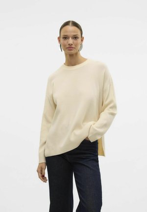 Cream-colored knit sweater with ribbed texture, round neck, and side slits, paired with dark blue wide-leg jeans.