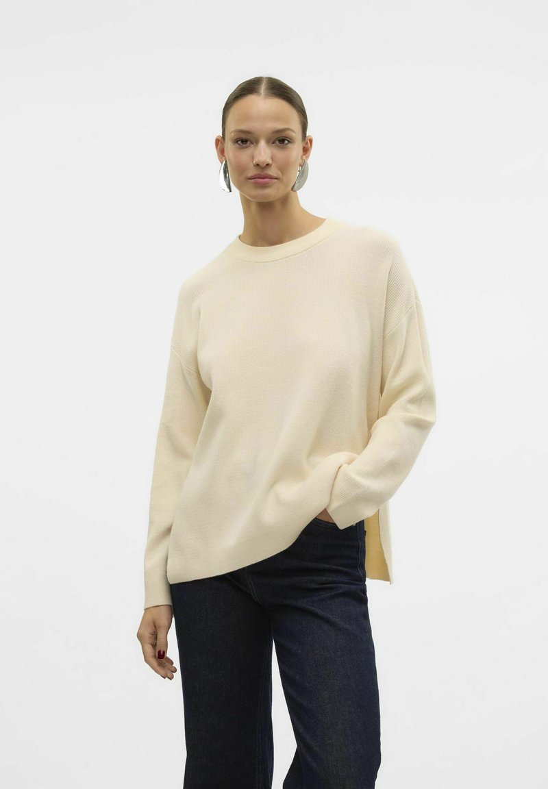 Cremefarvet striktrøje med ribbet struktur, rund hals og åbninger i siderne, parret med mørkeblå brede jeans.