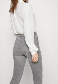 Blusa blanca de manga larga combinada con vaqueros de mezclilla gris ajustados. Los vaqueros cuentan con bolsillos traseros y un acabado texturizado.