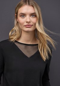 Młoda kobieta z długimi blond włosami, ubrana w czarną bluzkę z przezroczystym dekoltem, na jednolitym ciemnoszarym tle.