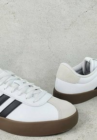 Biele kožené tenisky s šedými semišovými akcentmi, čiernymi bočnými pásmi a hnedými gumovými podrážkami. Navrhnuté s šnúrkami a zaobleným tvarom špičky.