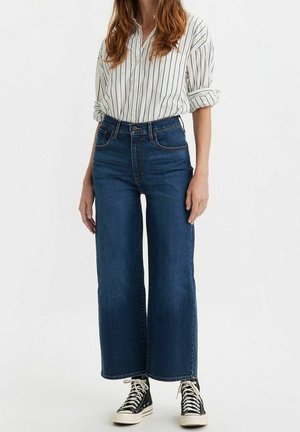 Vrouw draagt een wit gestreept blouse met knopen, een blauwe high waist wijde jeans en zwarte hoge sneakers met witte veters.