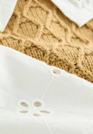Un tessuto bianco texturizzato con dettagli in pizzo floreale sovrappone un tessuto dorato lavorato a maglia con un motivo a diamante, mostrando un netto contrasto nei materiali.
