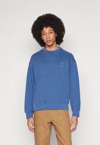 Sudadera azul con cuello redondo, puños y dobladillo acanalados, que presenta un pequeño parche de marca en el pecho. Corte clásico con una textura suave.