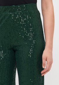 Roheline sädelevate pükste paar, millel on veniv vöökoht, tekstureeritud pind ja hõbedased detailid, mis püüavad valgust, kujutatud taljast kuni reideni.