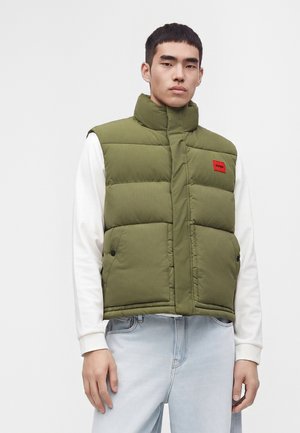 Giovane uomo che indossa un gilet imbottito verde con un patch logo rosso sopra una camicia bianca a maniche lunghe e jeans azzurri chiari su uno sfondo semplice.