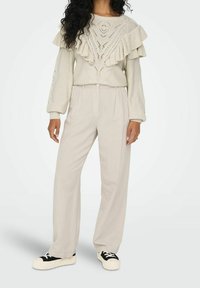 Sweter dzianinowy z falbaniastymi rękawami w jasnym kremowym kolorze, posiadający teksturowany wzór. Połączony z beżowymi spodniami o szerokich nogawkach i czarnymi butami w stylu sneakers.