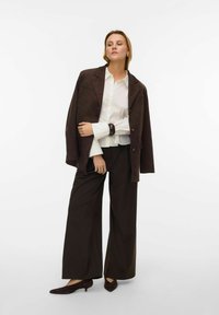 Brun ruskindsjakke i oversized model, hvid skjorte og mørkebrune bukser med vide ben. Fodtøjet er mørkebrune stiletter med spids tå. Enkel, ren design.