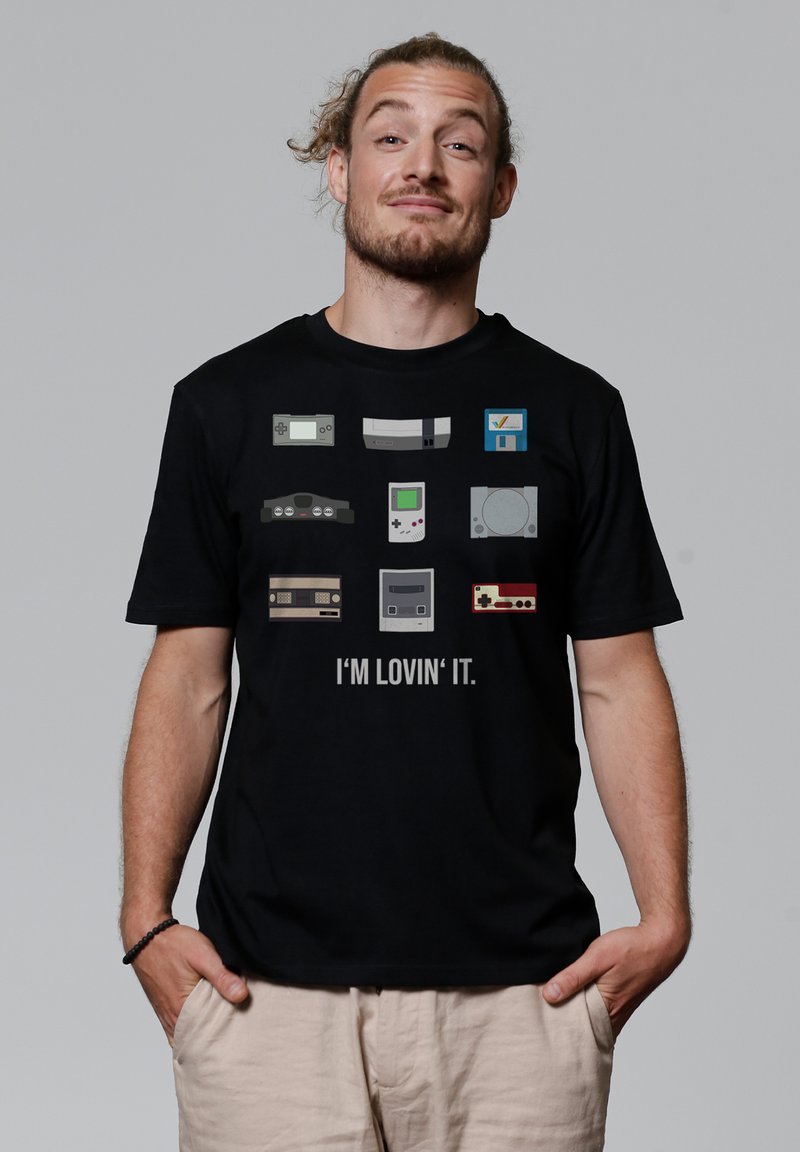 T-shirt noir avec des consoles de jeux pixelisées de différentes couleurs, avec le texte "JE L'AIME" affiché en dessous des designs.