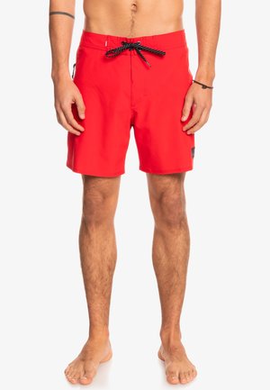 Red swim trunks made of quick-dry material, featuring a black drawstring waistband, side pocket, and an emblem on the leg.