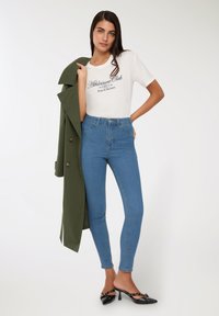 Biała koszulka z grafiką "Athleisure Club", zestawiona z niebieskimi dżinsami o wysokim stanie i oliwkowym płaszczem. Czarne clogs na obcasie.