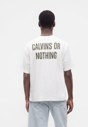 Mężczyzna odwrócony tyłem, ubrany w białą koszulkę z "CALVINS OR NOTHING" nadrukowanym na plecach pogrubionymi literami oraz jasnoniebieskie dżinsy.