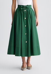 Gonna midi verde in cotone con vita aderente, grande fiocco, bottoni frontali e silhouette svasata. Abbinata a scarpe a punta con motivo.
