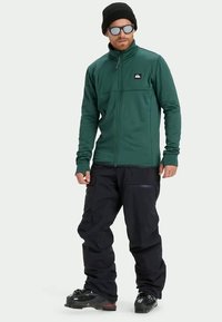 Tume roheline tõmblukuga fliisjakke kõrge kraega, rinnas logopatchiga, kantud mustade pükste ja lumisaabastega.