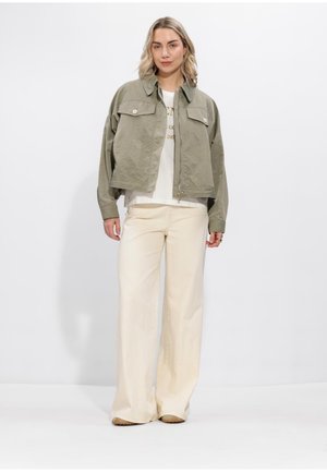 Vrouw staand met een licht olijfgroene jas, wit T-shirt met print en losse crèmekleurige broek tegen een effen witte achtergrond.