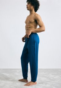 Rekbare blauwe sportieve broek met een elastische tailleband en zijzakken, met een subtiele logo op de linkerzijde, getoond op een model.