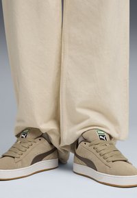 Beige suede sneakers with dark brown accents, white rubber soles, and tan laces. Wide, cream-colored pants drape over the shoes.