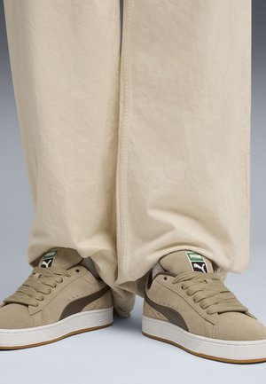 Beige suede sneakers with dark brown accents, white rubber soles, and tan laces. Wide, cream-colored pants drape over the shoes.