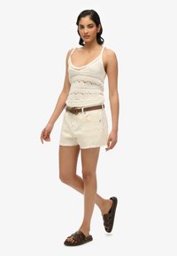 Top sin mangas de ganchillo blanco, pantalones cortos blancos desgastados, cinturón marrón y sandalias deslizantes marrón oscuro. Textura suave, diseño informal y ajuste relajado.