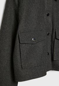 Poche de veste en laine gris foncé avec rabat et bouton argenté sur un fond de tissu texturé.