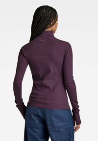 Lila ribbad långärmad tröja med hög krage och tumhål, med raglanärmar och en subtil logotyp på baksidan.