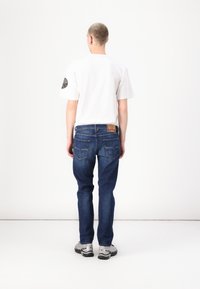 Blugi din denim albastru cu croială dreaptă și două buzunare din spate, purtați cu o cămașă albă cu mânecă scurtă, văzuți din spate. Adidași gri.