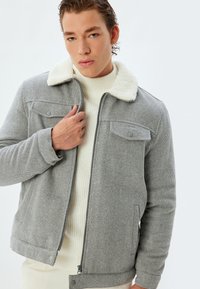 Grå hængebåndsjakke med en blød hvid krave, frontlynlås og skrå forlommer, båret over en cremet strikket sweater.