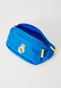 Plava tkanina torba s žutim patentnim zatvaračem, na prednjoj strani ima istaknut Real Madrid logo. Ima zaobljen oblik i mekanu teksturu.