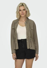 Ljust brunt knapplöst jacka med dragsko i midjan, bröstfickor och relaxed fit, kombinerat med svarta denimshorts.