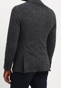 Uomo con un blazer testurizzato grigio scuro e pantaloni scuri, visto da dietro su uno sfondo chiaro e semplice.