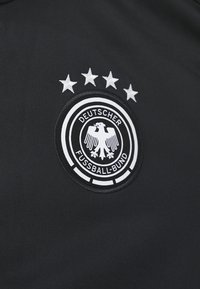 Μαύρο ύφασμα με εγ embossing κυκλικό σήμα. Το σήμα διαθέτει έναν αετό, κείμενο "DEUTSCHER FUSSBALL-BUND" και πέντε λευκά αστέρια από πάνω.