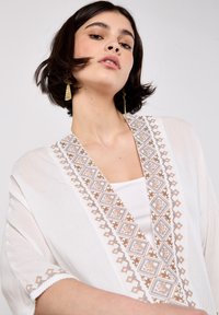 White kimono with a geometric brown and light blue embroidered pattern along the neckline, featuring a lightweight, textured fabric and wide sleeves.