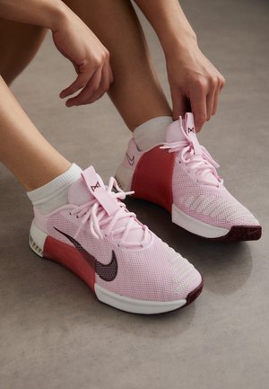 Osoba sznurowa różowe buty do biegania Nike z białymi podeszwami i bordowymi akcentami, siedząc na gładkiej szarej podłodze.