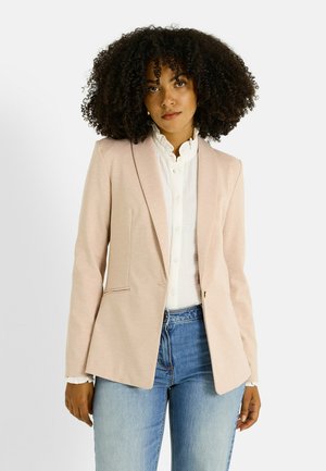 Blazer rosa claro con hombros sin estructura, solapa con muesca y dos bolsillos delanteros, llevado sobre una camisa blanca de botones y vaqueros azules.