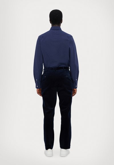 Mørkeblå skjorte med knapper og hvite prikker, kombinert med mørke cordbukser. Hvit sneakers fullfører looket. Utsikt fra baksiden.