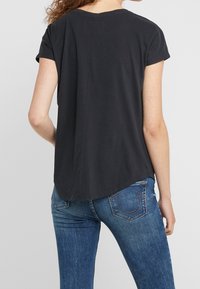 Woman with curly hair wearing a black short-sleeve t-shirt and blue denim jeans, shown from the back against a plain background.