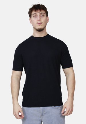 Giovane uomo con capelli corti e scuri, che indossa una maglietta nera a maniche corte semplice e jeans azzurri, in piedi contro uno sfondo chiaro.