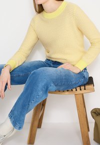 Femme assise sur un tabouret en bois avec un siège noir tissé, portant un pull texturé jaune, un jean bleu, des chaussettes blanches et des chaussures argentées.