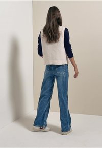 Beige stickad väst över en marinblå långärmad topp, kombinerad med blå vida jeans och vita sneakers. Slät textur, avslappnad passform.