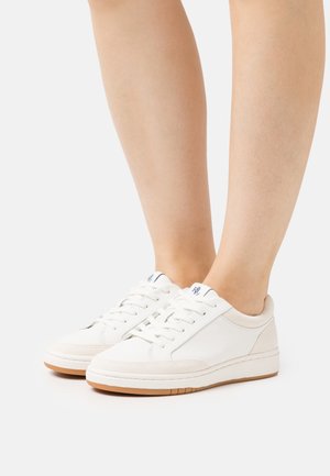 Personne portant des baskets blanches en cuir avec des semelles en gomme et des lacets blancs, debout sur un fond clair uni.