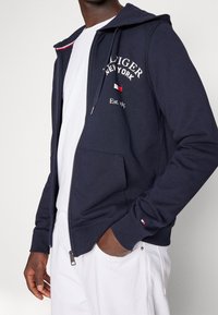 Sudadera con capucha con cremallera en azul marino, fabricada con tela suave, que presenta un estampado de logo, costuras en contraste y un bolsillo frontal. La capucha tiene un cordón ajustable y puños acanalados.