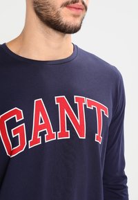 Laivastonsininen pitkähihainen T-paita puuvillasta, jossa on punainen "GANT"-logo rinnassa. Pyöreä kaula-aukko ja pehmeä tekstuuri, minimalismi suunnittelussa.