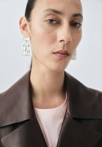 Brązowy skórzany płaszcz z dużymi klapami, noszony na jasnoróżowej bluzce. Duże, przezroczyste, geometryczne kolczyki dodają wyrazistego akcentu. Gładka tekstura skóry.