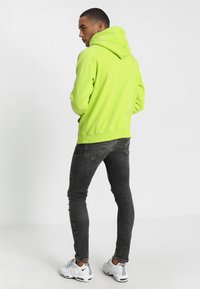 Sudadera verde neón con capucha ajustable, puños acanalados y dobladillo, combinada con unos jeans ajustados negros desgastados y zapatillas deportivas blancas.