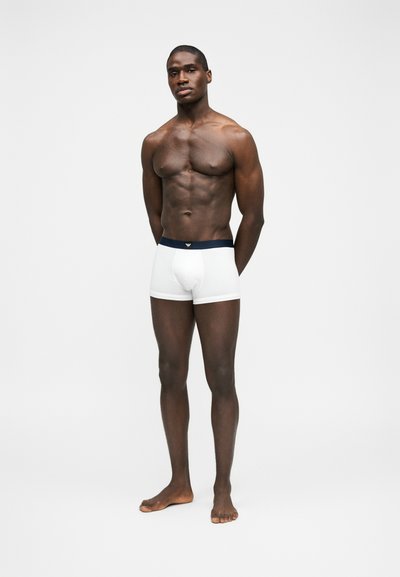 Μυώδης άνδρας στέκεται ξυπόλητος μπροστά σε λευκό φόντο, φορώντας λευκά μπόξερ με σκούρα μέση, με τα χέρια πίσω από την πλάτη, κοιτάζοντας μπροστά.