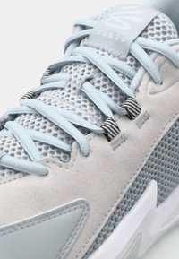 Basket de sport gris clair avec une empeigne en mesh, des accents en daim lisse et des lacets bleu clair. Présente une semelle texturée et une languette rembourrée.