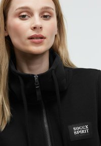 Black zip-up jacket with a high collar and drawstring. Features a logo patch on the chest and textured zippers. Soft, smooth fabric.