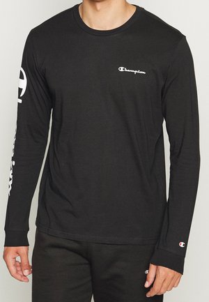 Homme portant un t-shirt noir à manches longues Champion avec logo blanc sur la poitrine, la manche gauche et le poignet gauche, ainsi qu'un pantalon noir assorti avec logo.