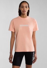 Hellrosa Baumwoll-T-Shirt mit kurzen Ärmeln, mit einem weißen gedruckten Logo auf der Vorderseite. Kombiniert mit schwarzen, eng anliegenden Shorts.